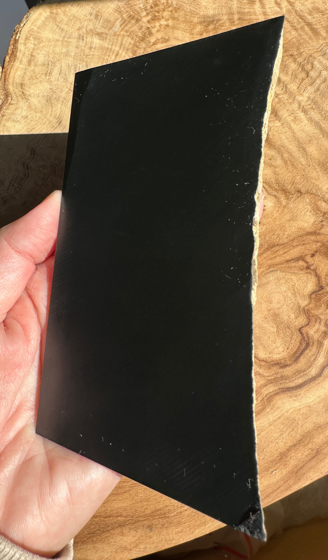 A hand holding a thick, solid black jade rough, natural wood edge, set against a rustic wooden background.