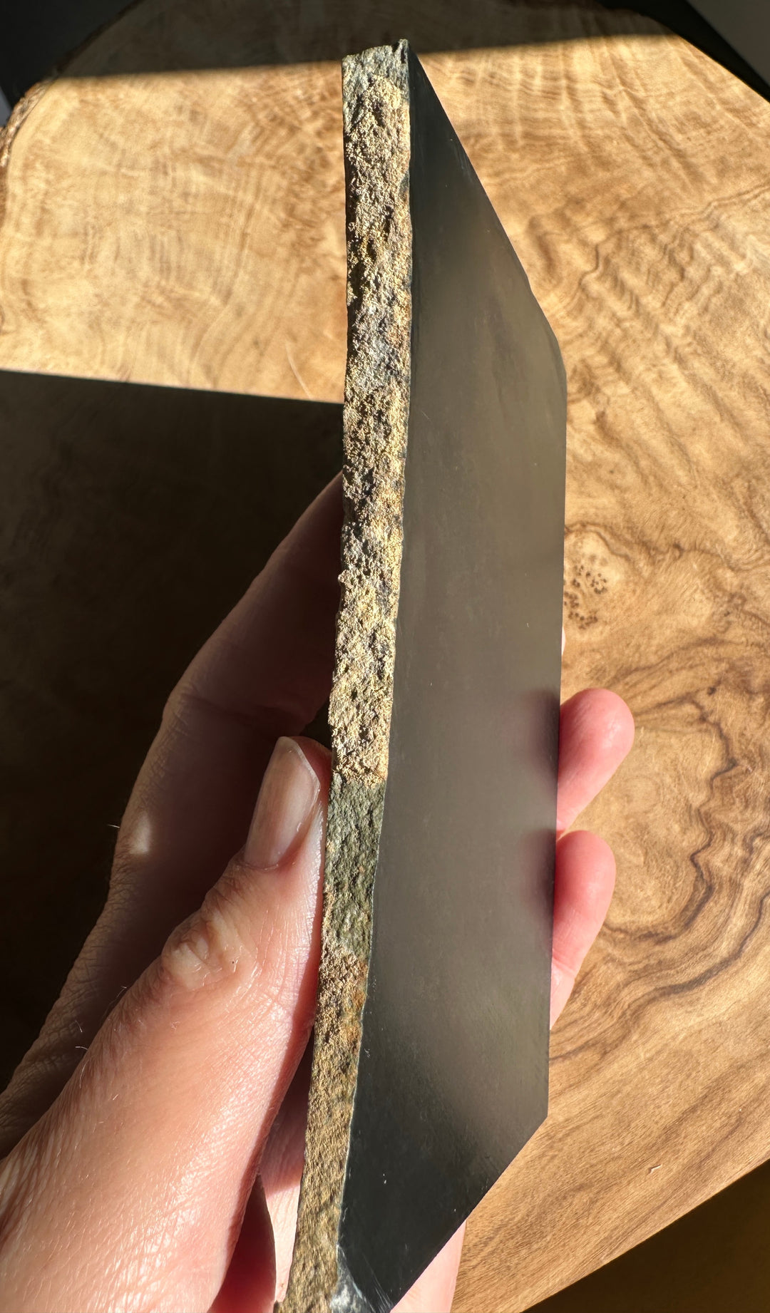 A hand holding a rectangular dark slab, showing its rough textured natural edge, against a warm wood-grain background.