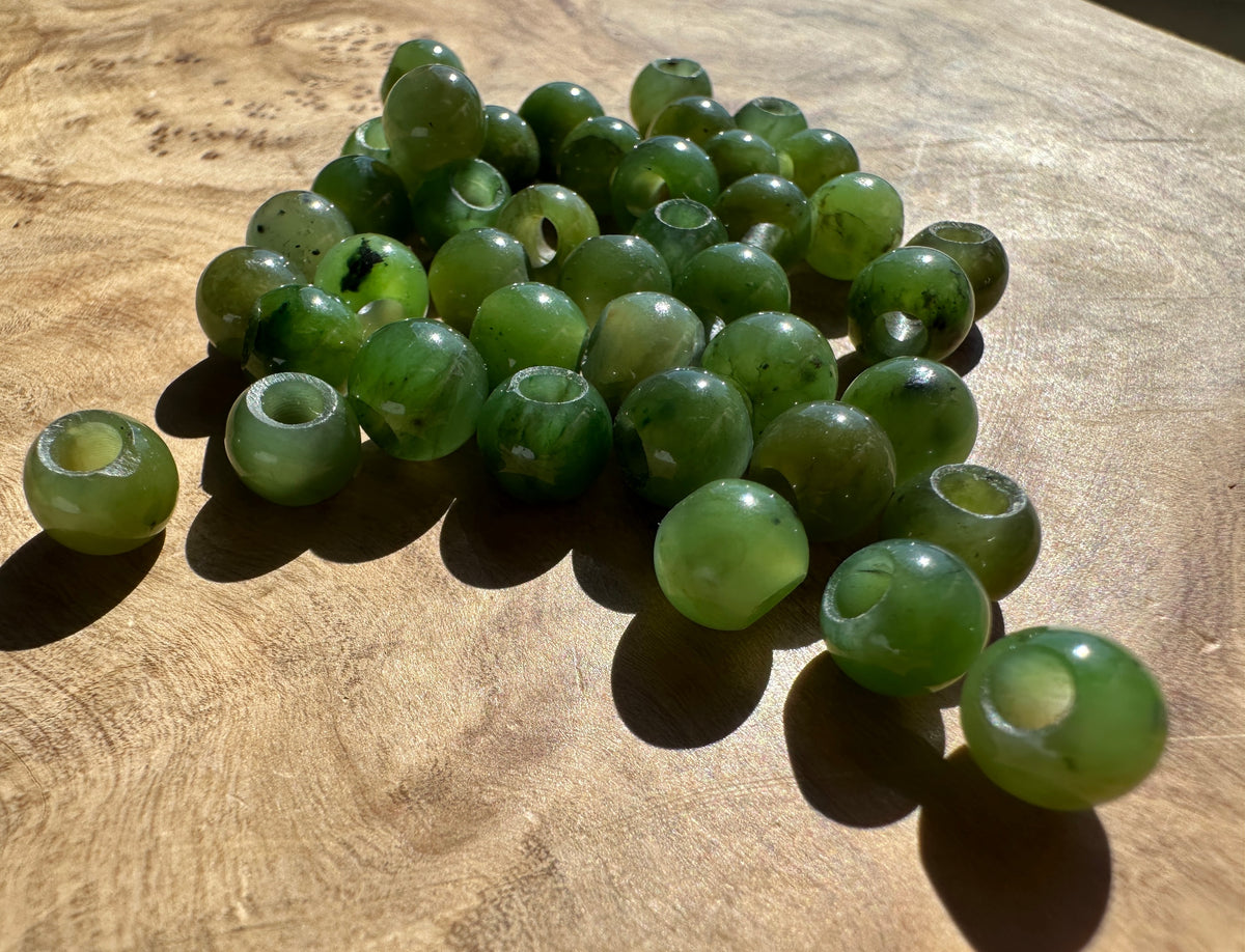 A pile of green translucent jade beads with hollow centers, resting on a textured wooden surface under direct sunlight.