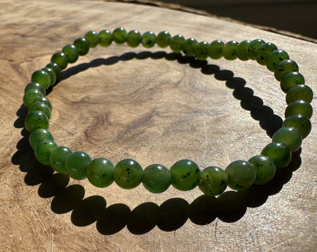 A green beaded stretch bracelet made of polished round beads with dark inclusions, displayed on a wooden surface under direct sunlight.