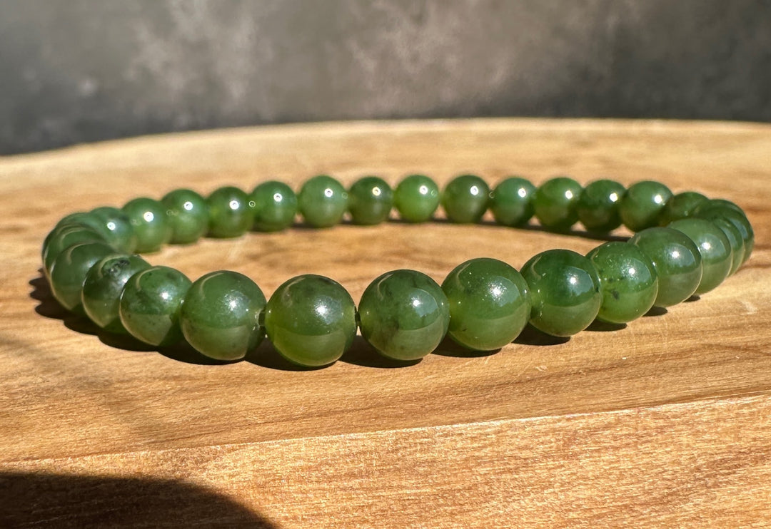 A green beaded bracelet made of polished round stones, displayed on a wooden surface with natural sunlight and shadows.