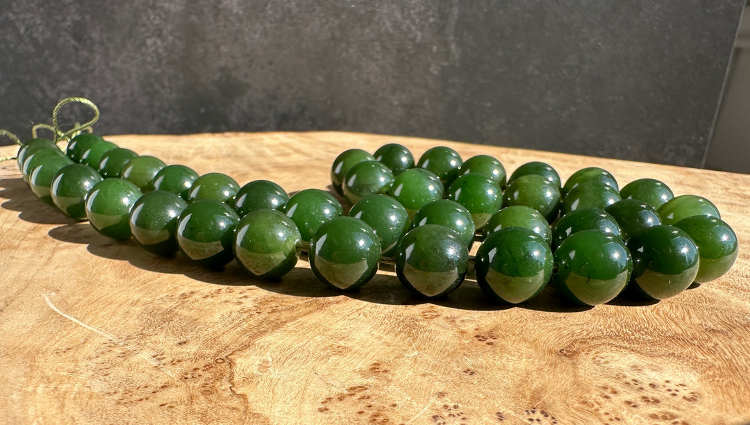 A strand of polished dark green jade-like beads arranged in a curve on a natural wood surface, with a dark gray textured wall in the background.