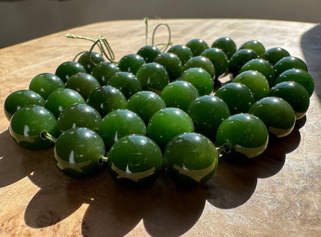 A group of polished dark green jade round beads strung on thin cords, arranged on a wooden surface in direct sunlight.