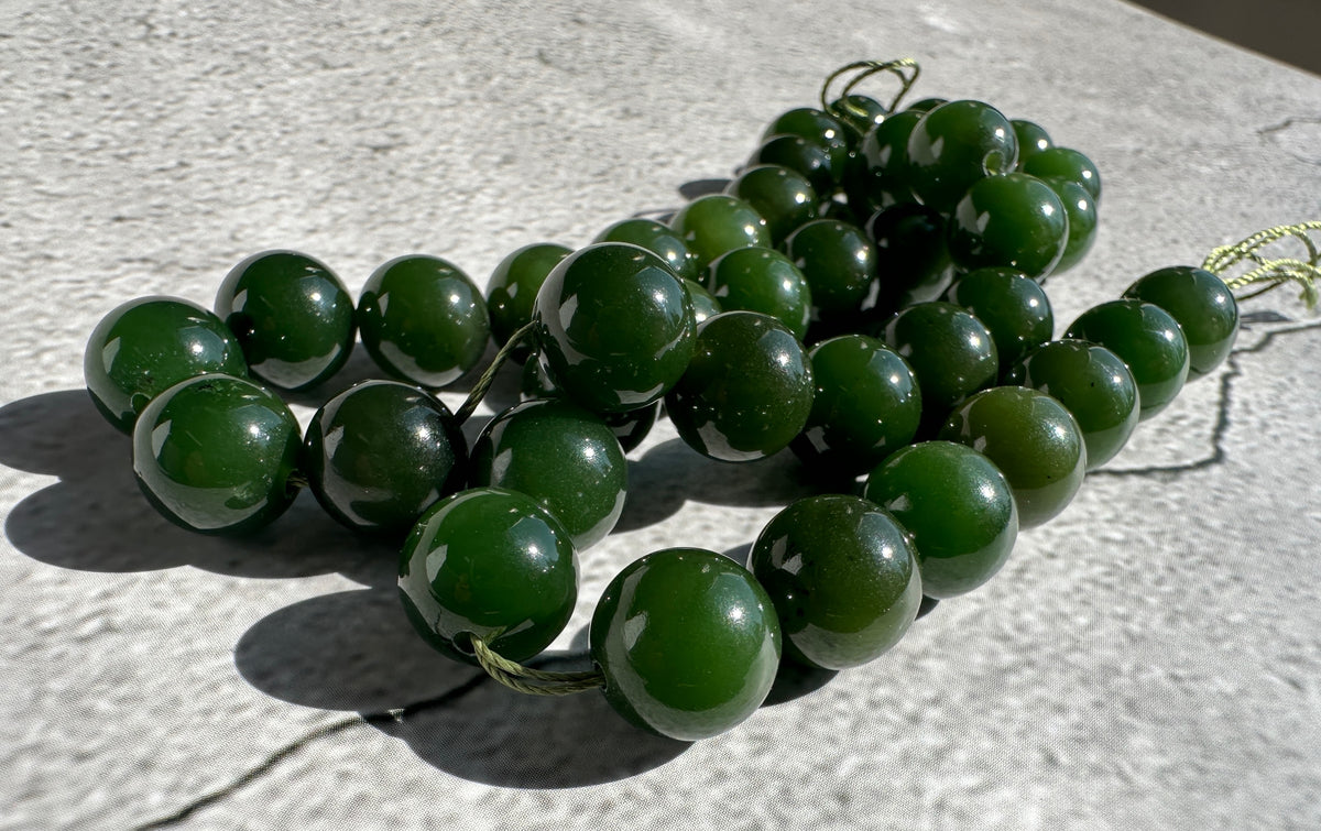 A strand of polished dark green jade beads strung on a thin cord, resting on a light gray textured surface in bright sunlight.