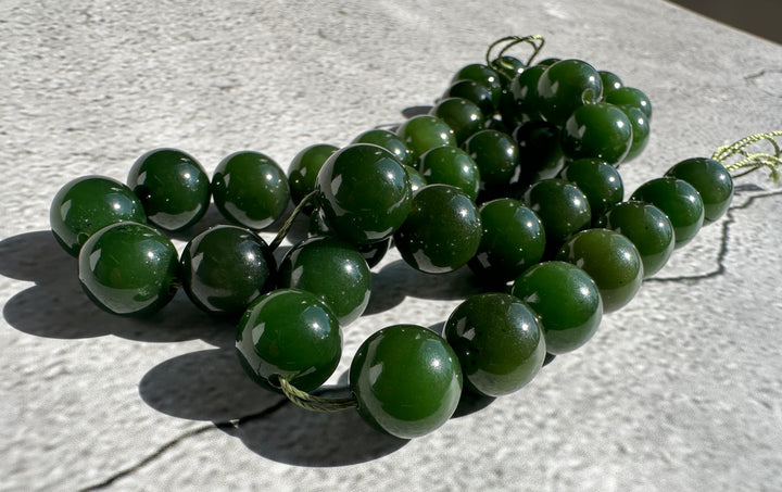 A strand of polished dark green jade beads strung on a thin cord, resting on a light gray textured surface in bright sunlight.