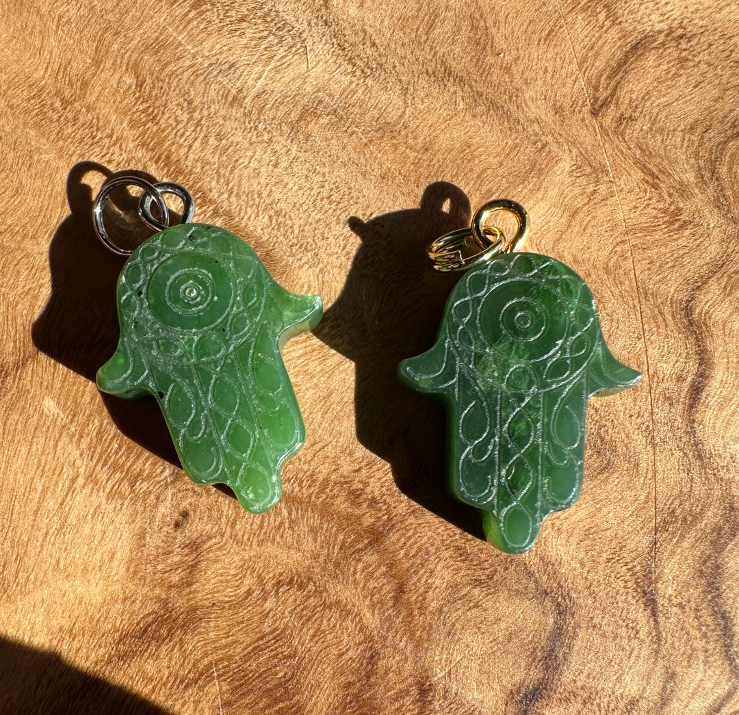 Two green carved hamsa hand pendants with intricate filigree patterns, one with a silver ring and the other with a gold ring, displayed on a textured wooden surface.