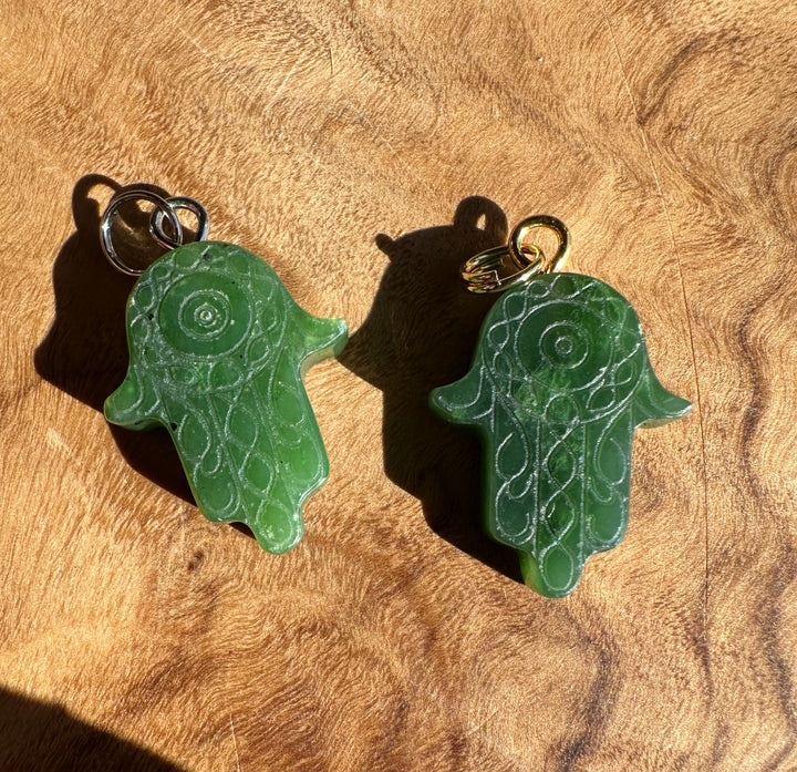 Two green carved hamsa hand pendants with intricate filigree patterns, one with a silver ring and the other with a gold ring, displayed on a textured wooden surface.