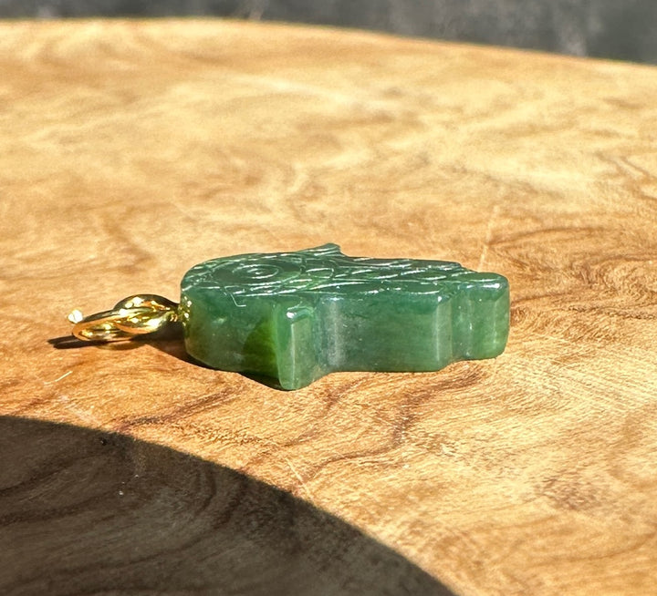 A green jade pendant with a gold bail, resting on a textured wooden surface.