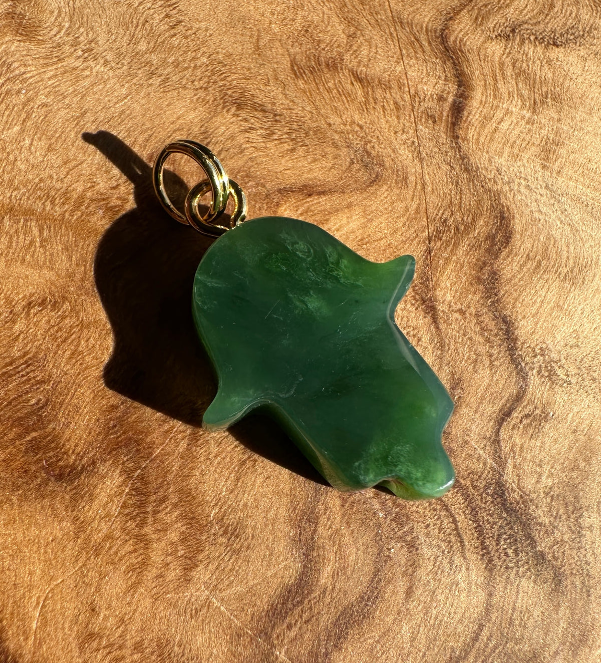 A green hamsa hand pendant with a gold-tone bail, resting on a textured brown wood surface.