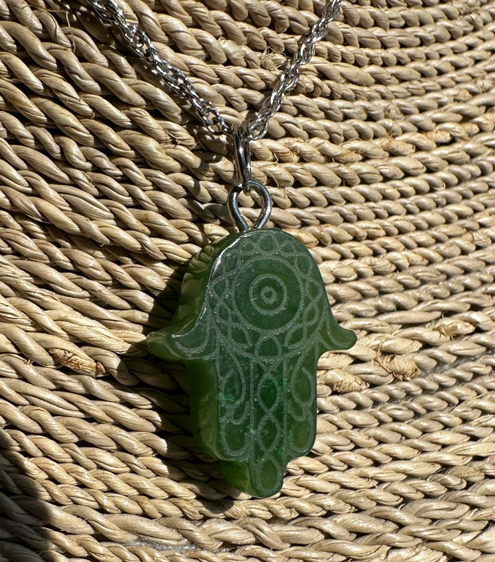 A green hamsa hand pendant necklace with carved circular patterns, hanging from a silver chain on a woven straw background.