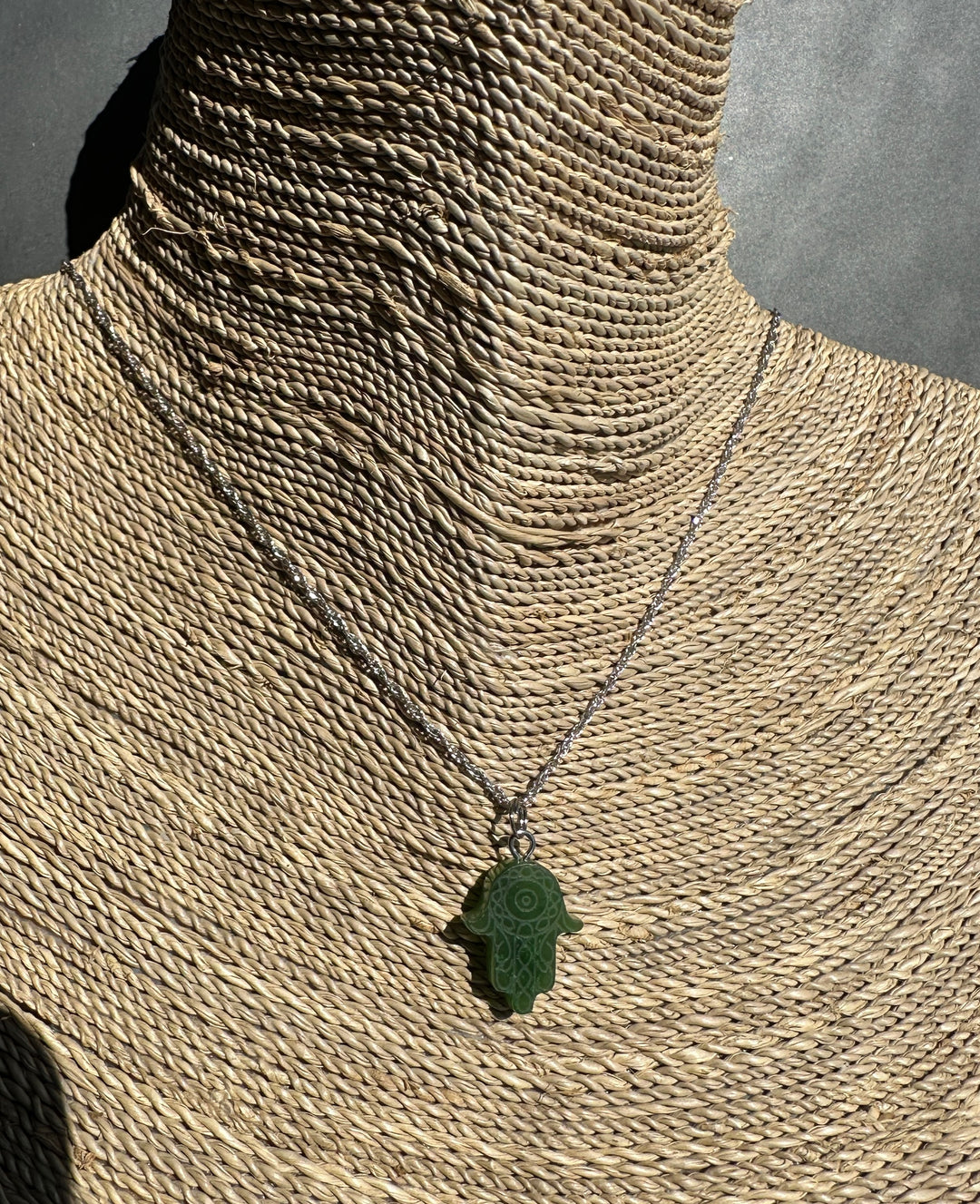 A dark green carved Hamsa hand pendant necklace on a silver chain, displayed on a woven straw mannequin neck against a gray background.