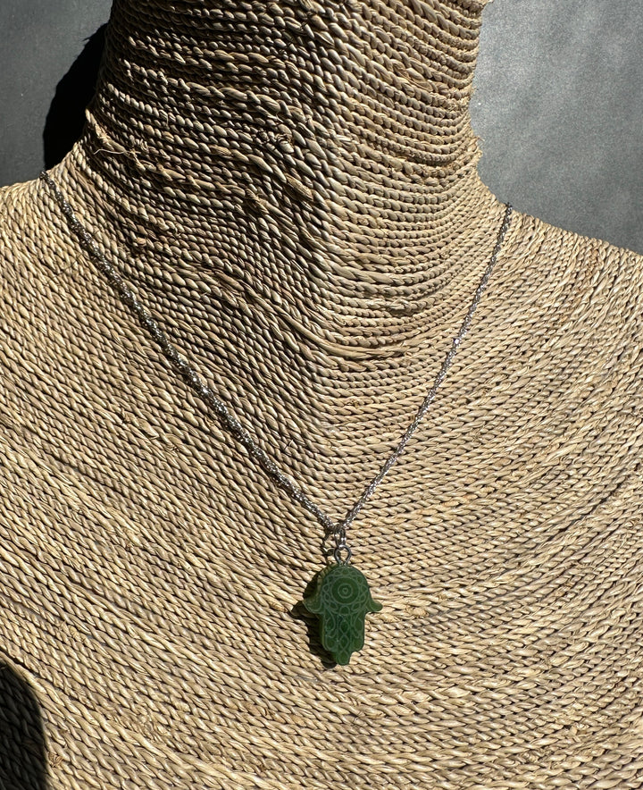 A dark green carved Hamsa hand pendant necklace on a silver chain, displayed on a woven straw mannequin neck against a gray background.