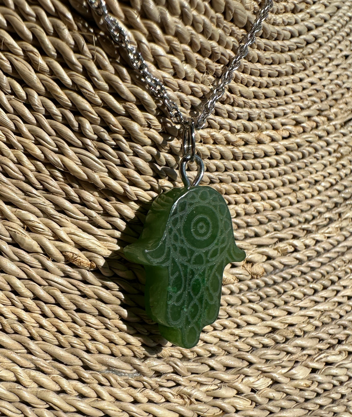 A green hamsa hand pendant necklace with a silver-tone chain, displayed on a woven straw surface.