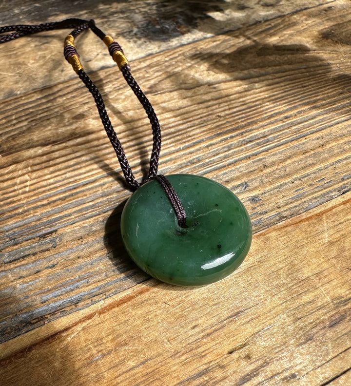 Green jade donut pendant on dark brown braided nylon cord resting on a wooden surface.