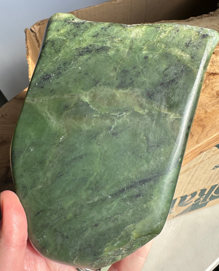 A hand holding a large, polished green jade with natural dark green veining, set against a cardboard box background.