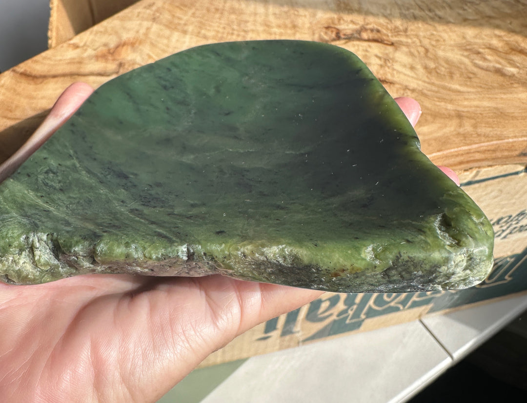 A hand holding a large, polished green stone or jade-like slab with natural dark veining, set against a wooden box background.