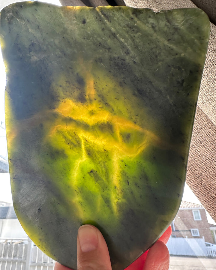 A hand holding a large, irregularly shaped polished jade with natural marbling. The stone features a dominant dark green body with prominent bright yellow-green and yellow veins, and dark gray/black inclusions.