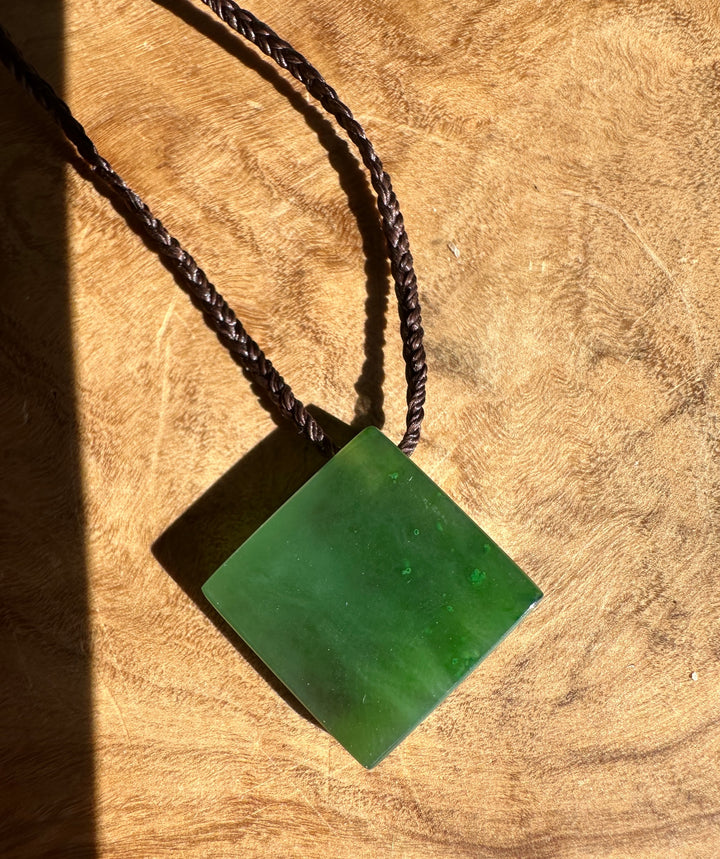 A square green jade pendant necklace on a dark braided cord, displayed on a textured wooden surface with a strong diagonal shadow.