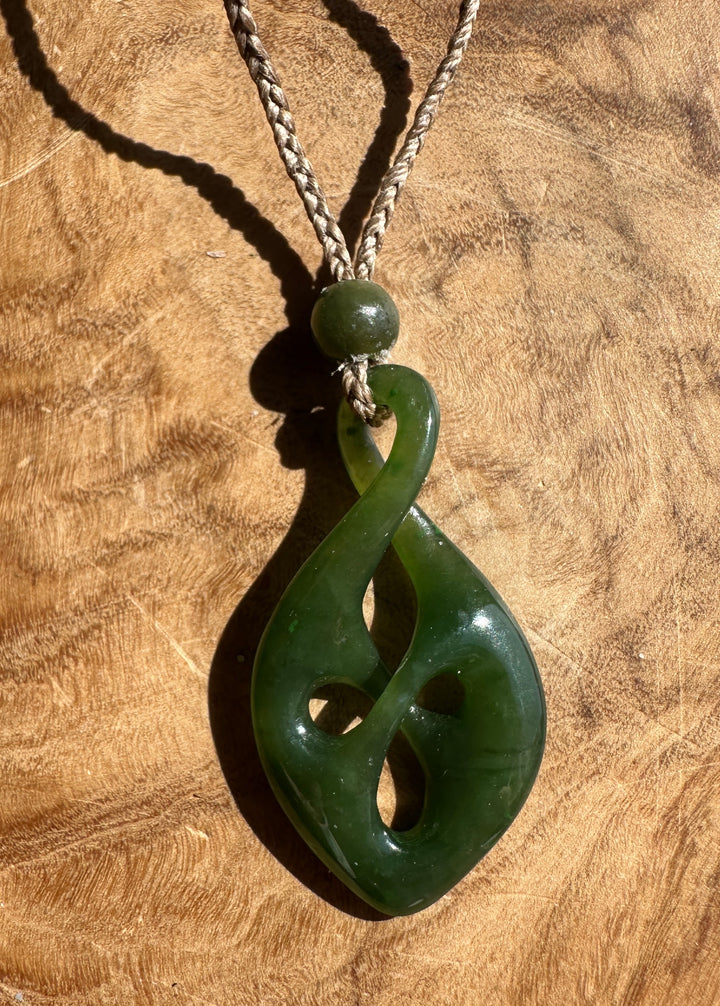 A carved green jade pendant necklace with a braided cord, displayed on a textured wooden surface.