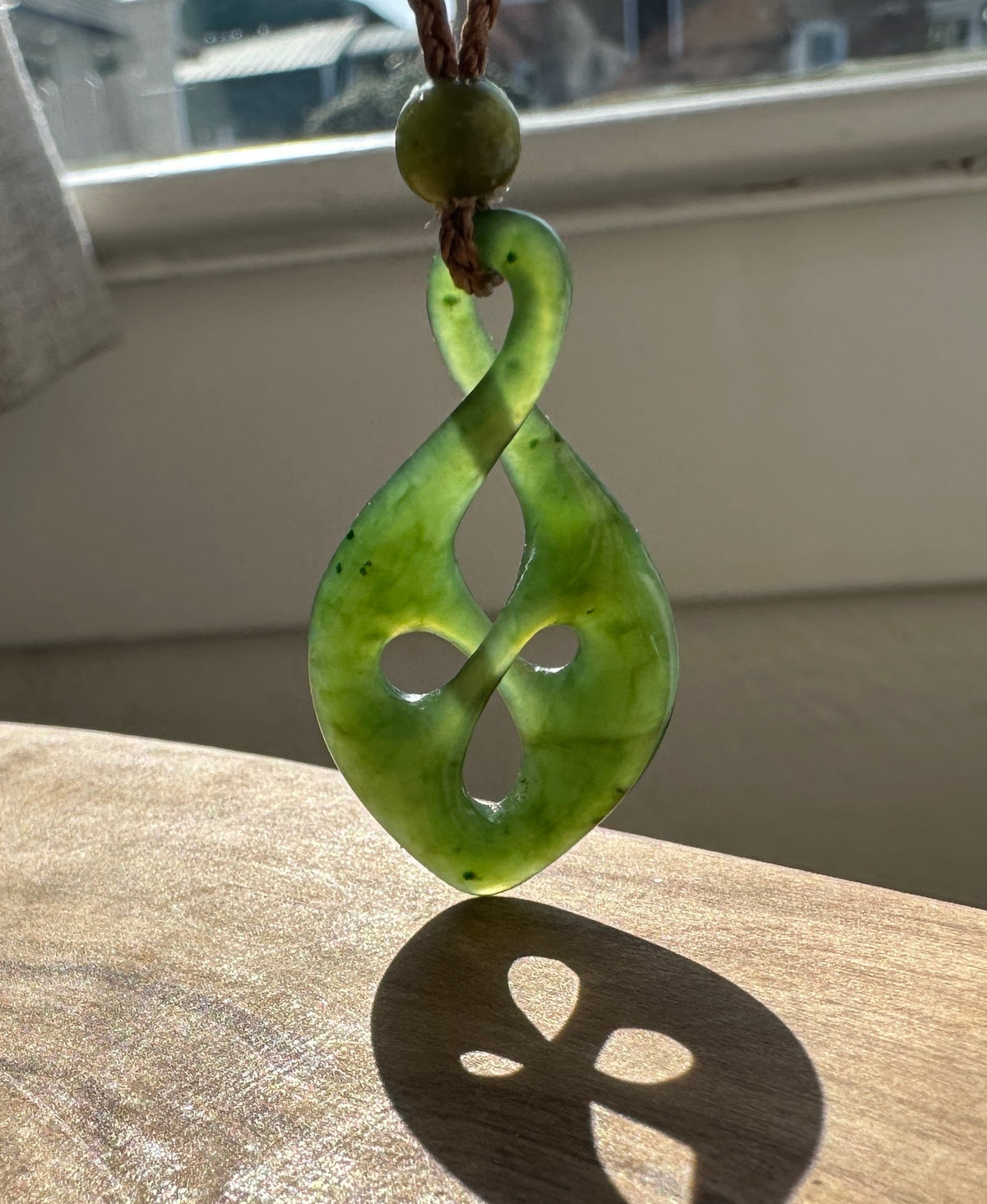 A green carved pendant with a knot-like design hangs from a brown cord and bead, resting on a wooden surface near a window.