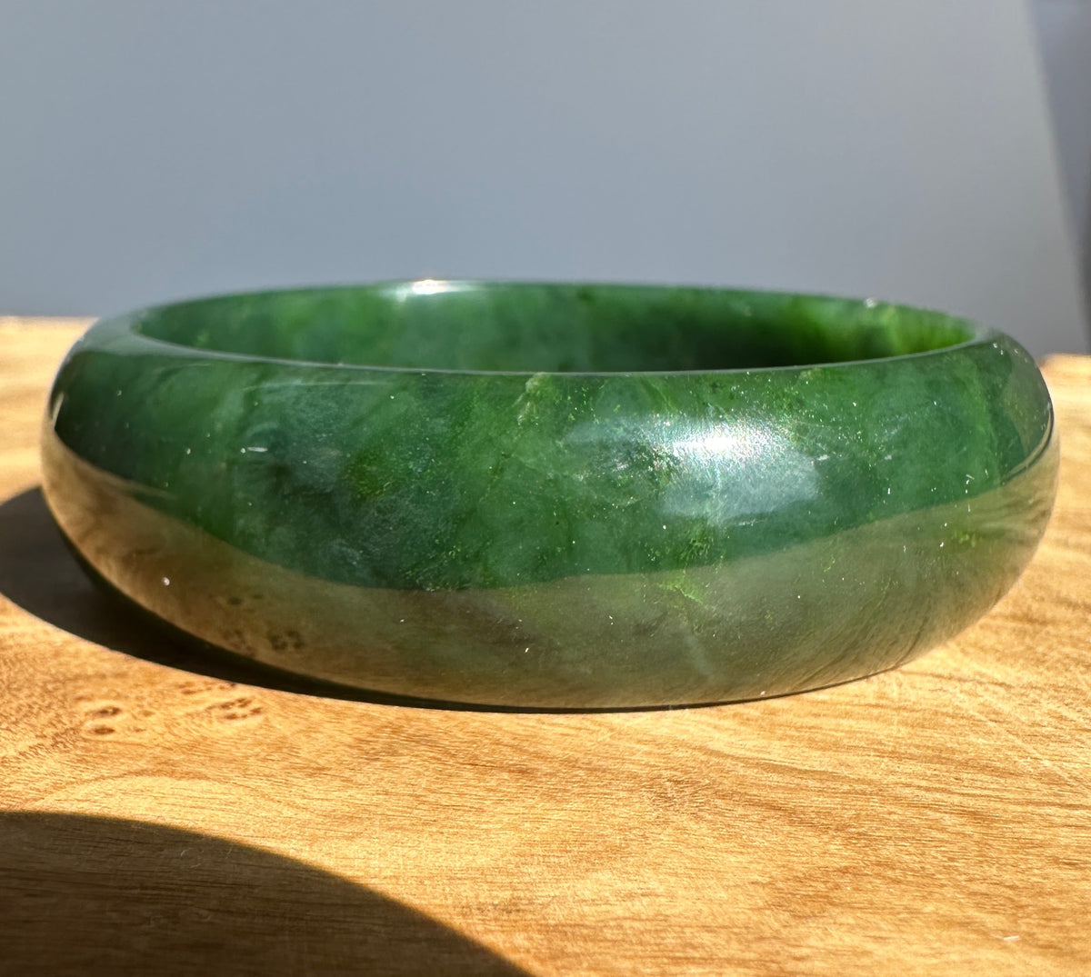 A polished green jade bangle bracelet resting on a wooden surface under direct sunlight.