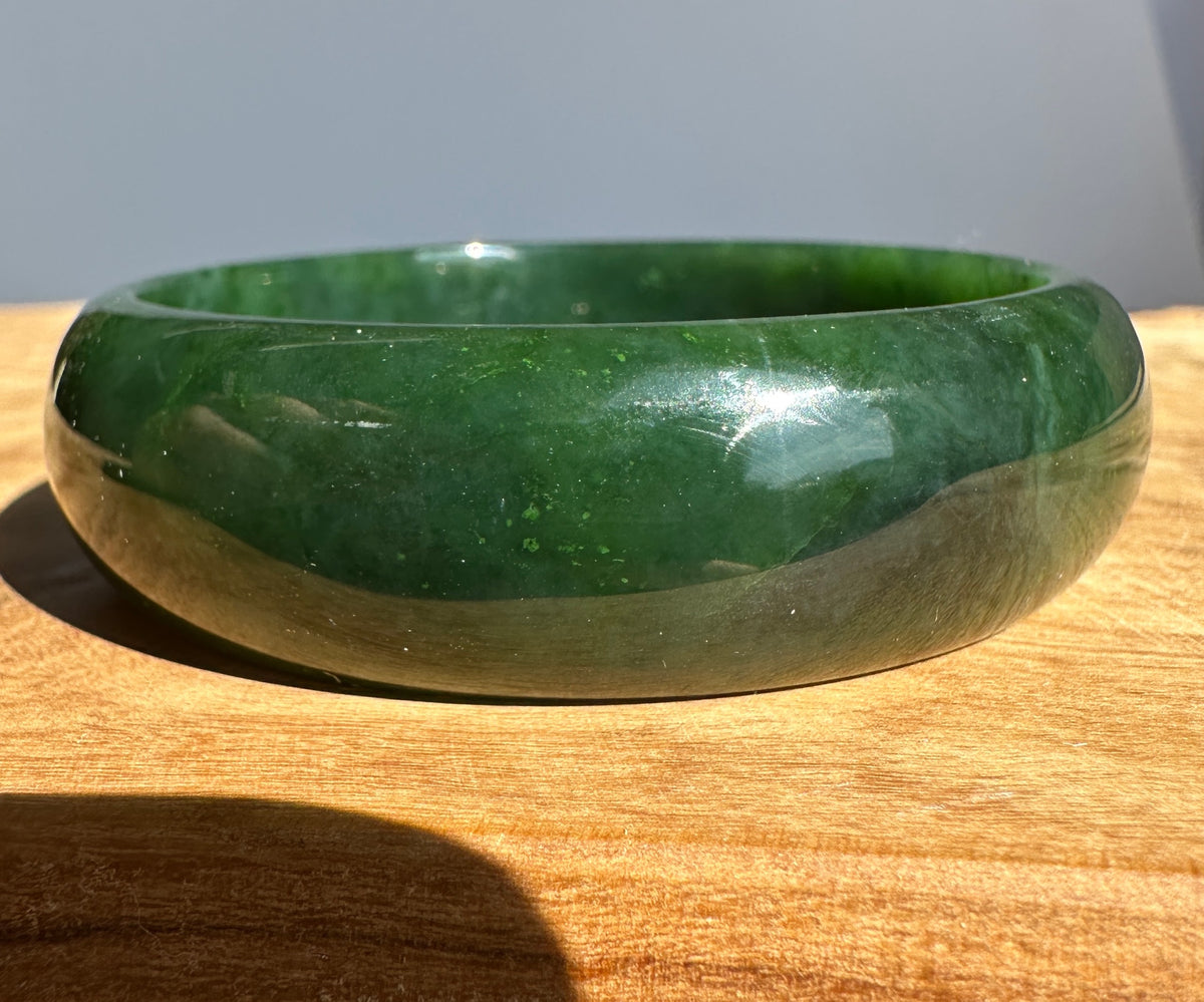 A polished dark green jade bangle bracelet resting on a wooden surface with a neutral gray background.