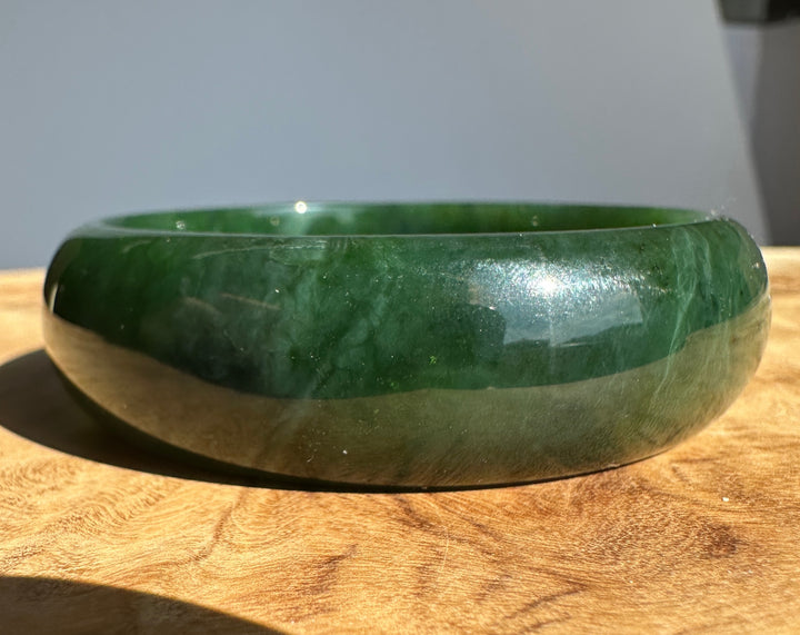 A polished dark green jade bangle bracelet resting on a wooden surface against a neutral gray background.