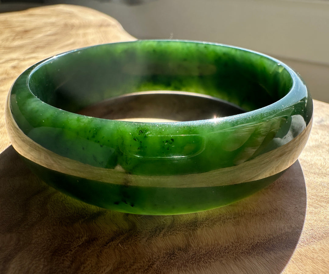 A polished green jade bangle bracelet resting on a wooden surface, showing vibrant green and lighter marbled patterns.