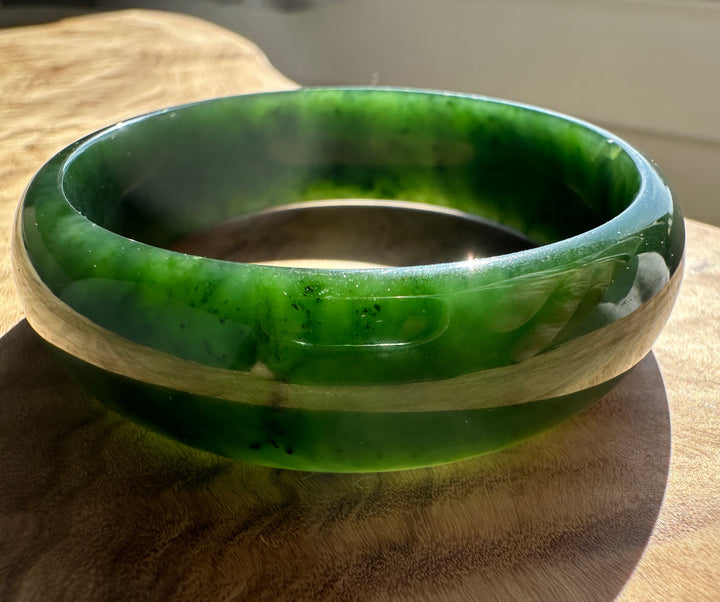 A polished green jade bangle bracelet resting on a wooden surface, showing vibrant green and lighter marbled patterns.