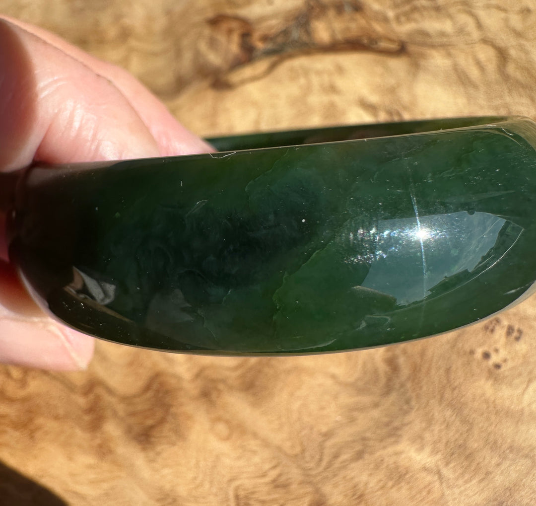 A close-up of a hand holding a polished dark green jade bangle against a natural wood grain background.