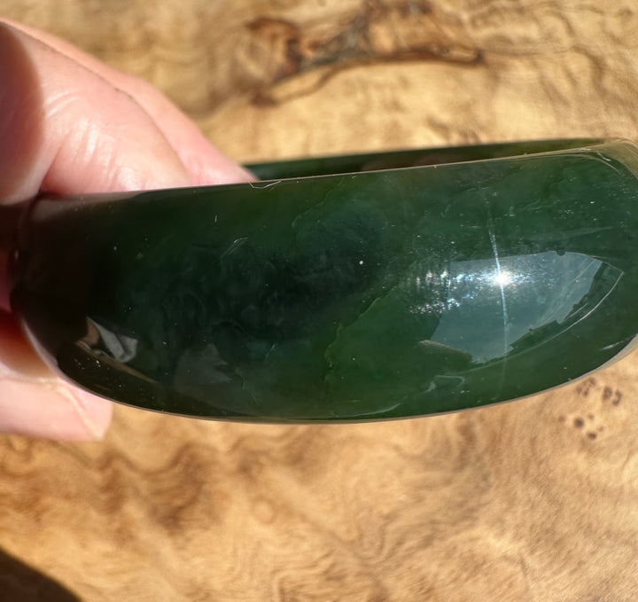 A close-up of a hand holding a polished dark green jade bangle against a natural wood grain background.