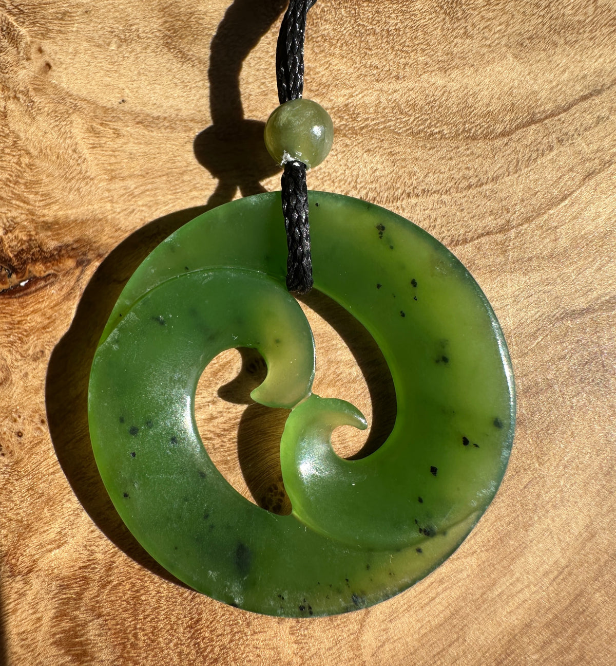 A green carved stone pendant necklace with a black braided cord, displayed on a textured wooden surface.