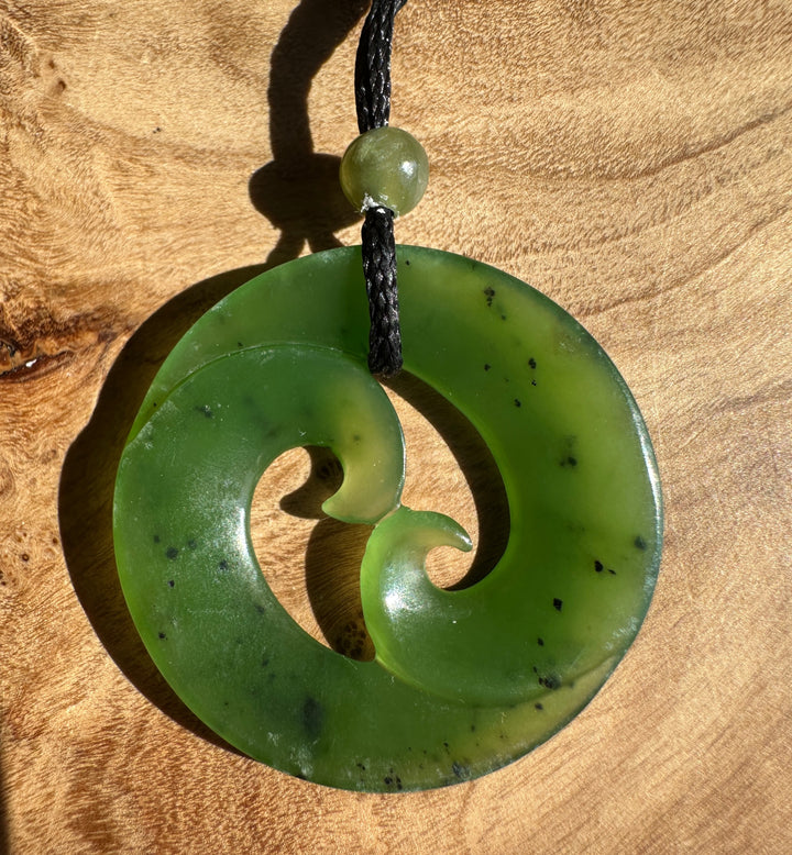 A green carved stone pendant necklace with a black braided cord, displayed on a textured wooden surface.