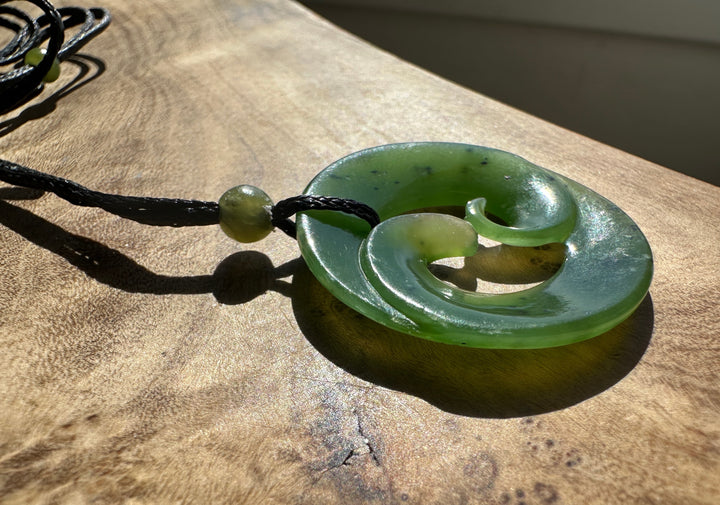 A green carved pendant necklace with a black cord, resting on a rustic wooden surface under direct sunlight.