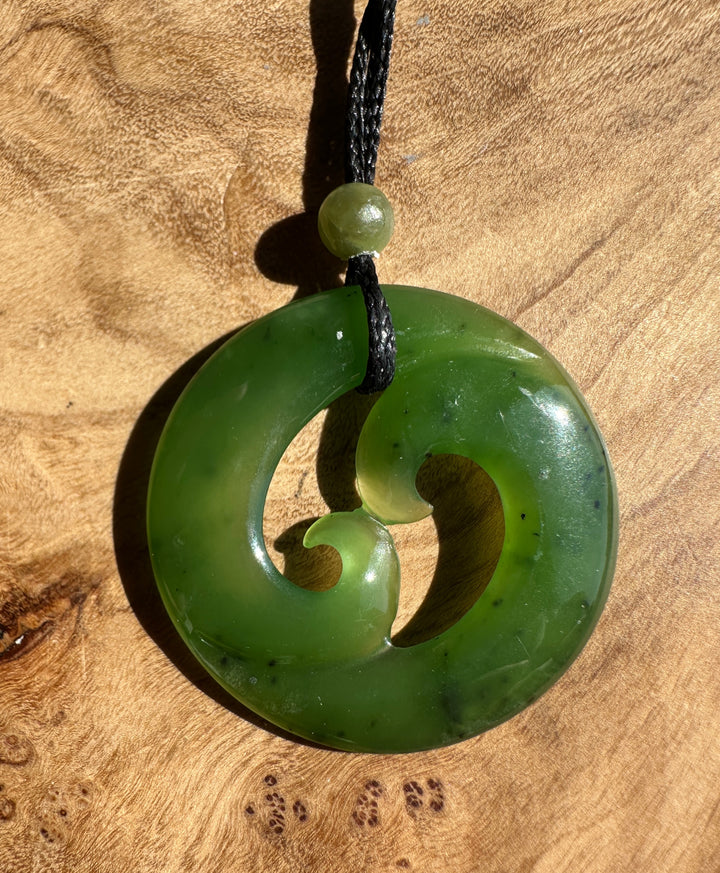 A green stone pendant necklace with a carved spiral design, suspended from a black cord with a small green bead, displayed on a textured wooden surface.