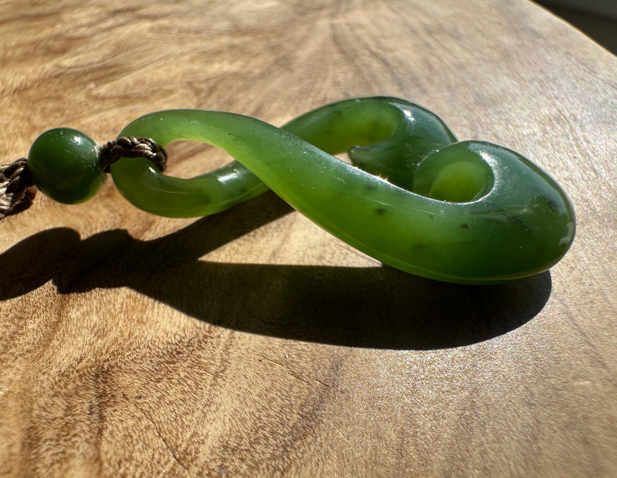 A green jade pendant with a curved, looped design and a small green bead on a cord, resting on a wooden surface.