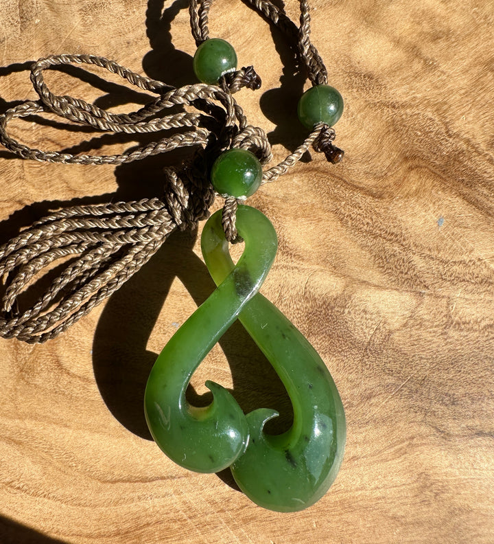 A green jade pendant necklace with a carved hook design, suspended from a braided brown cord with green beads, displayed on a wooden surface.