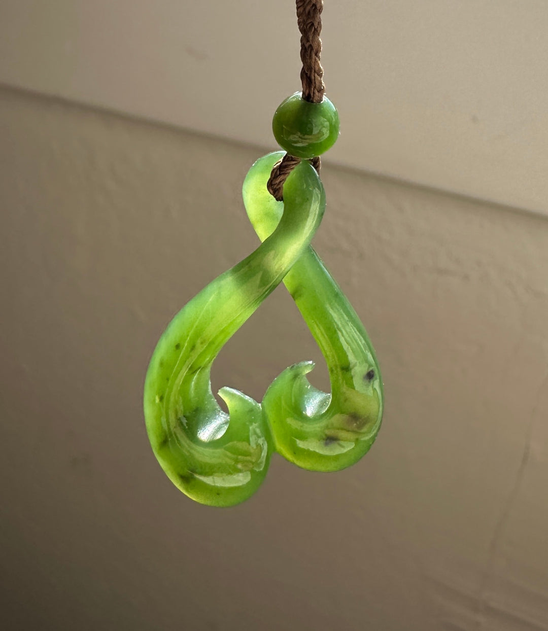A green jade pendant with a twisted, curved design and a small green bead at the top, hanging from a brown cord against a beige wall.
