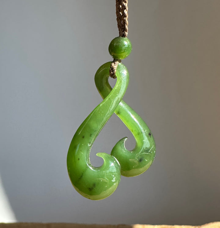 A green polished carved pendant or necklace charm with a curved hook-like shape, hanging from a brown cord against a soft gray background.