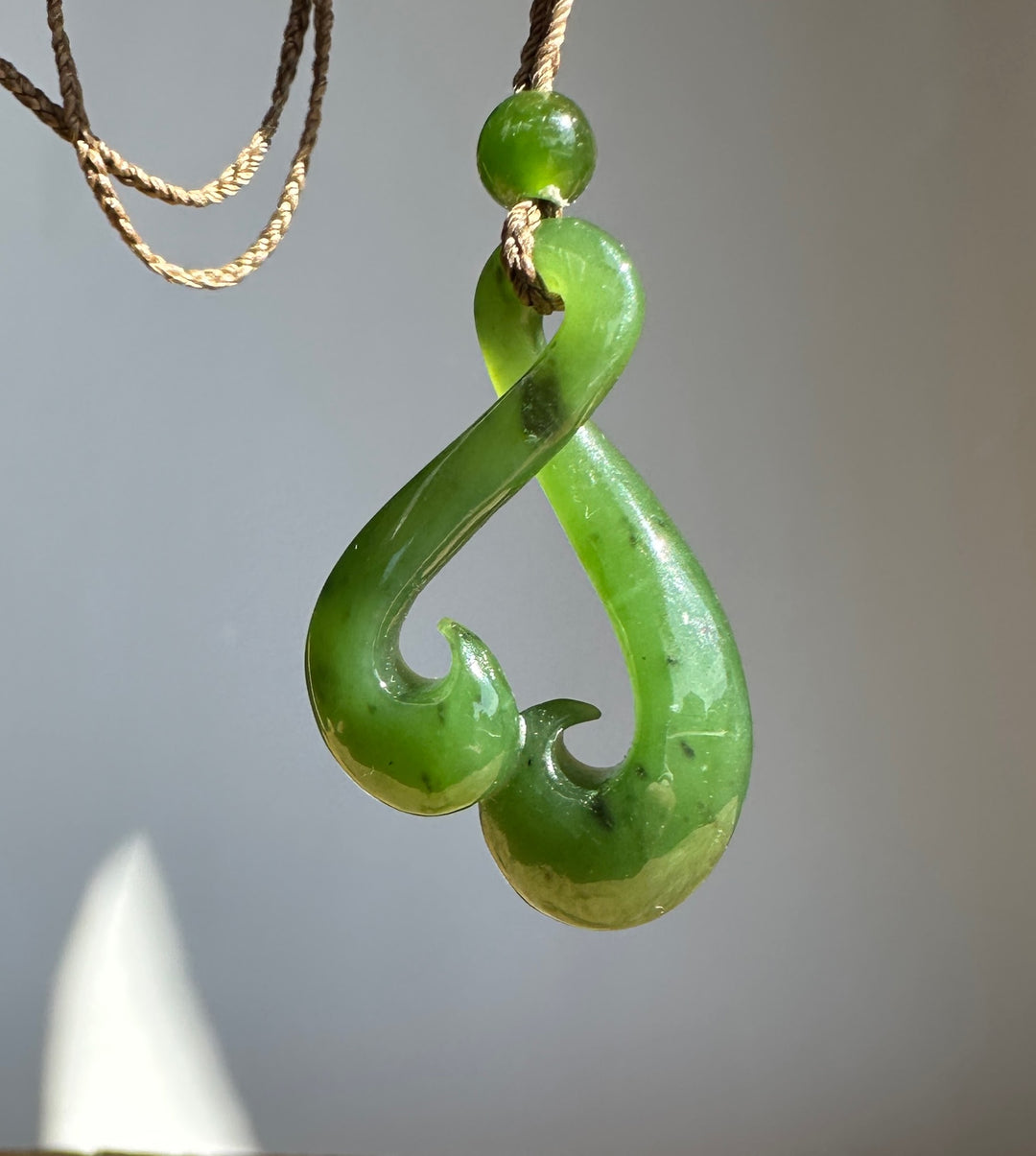 A green jade pendant shaped like a hook, suspended from a tan cord necklace against a neutral gray background.