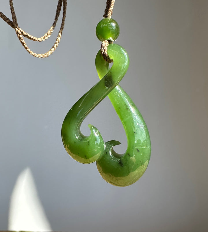 A green jade pendant shaped like a hook, suspended from a tan cord necklace against a neutral gray background.