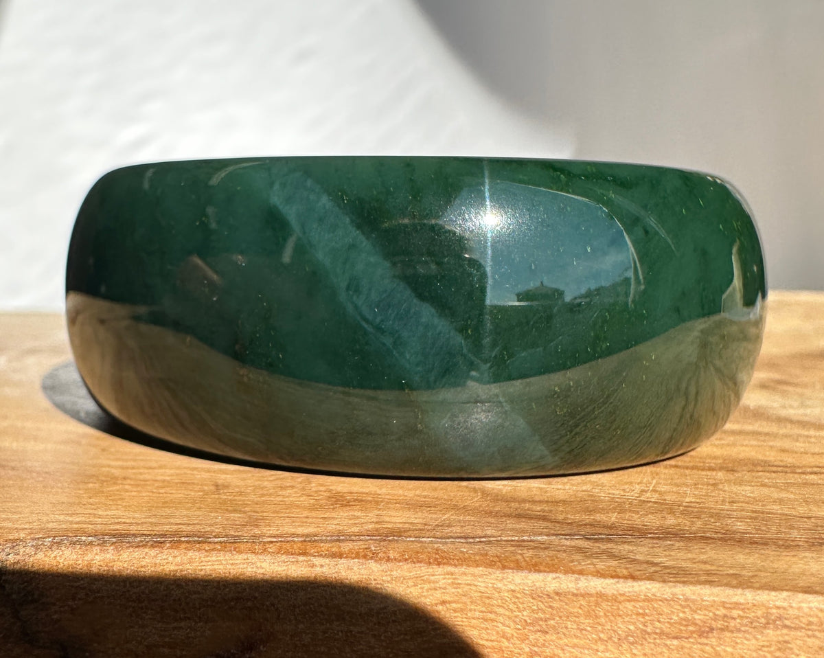 A polished dark green stone bangle bracelet resting on a wooden surface with a white background.