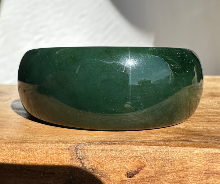 A polished dark green jade bangle with a smooth, rounded shape, resting on a wooden surface under direct sunlight.