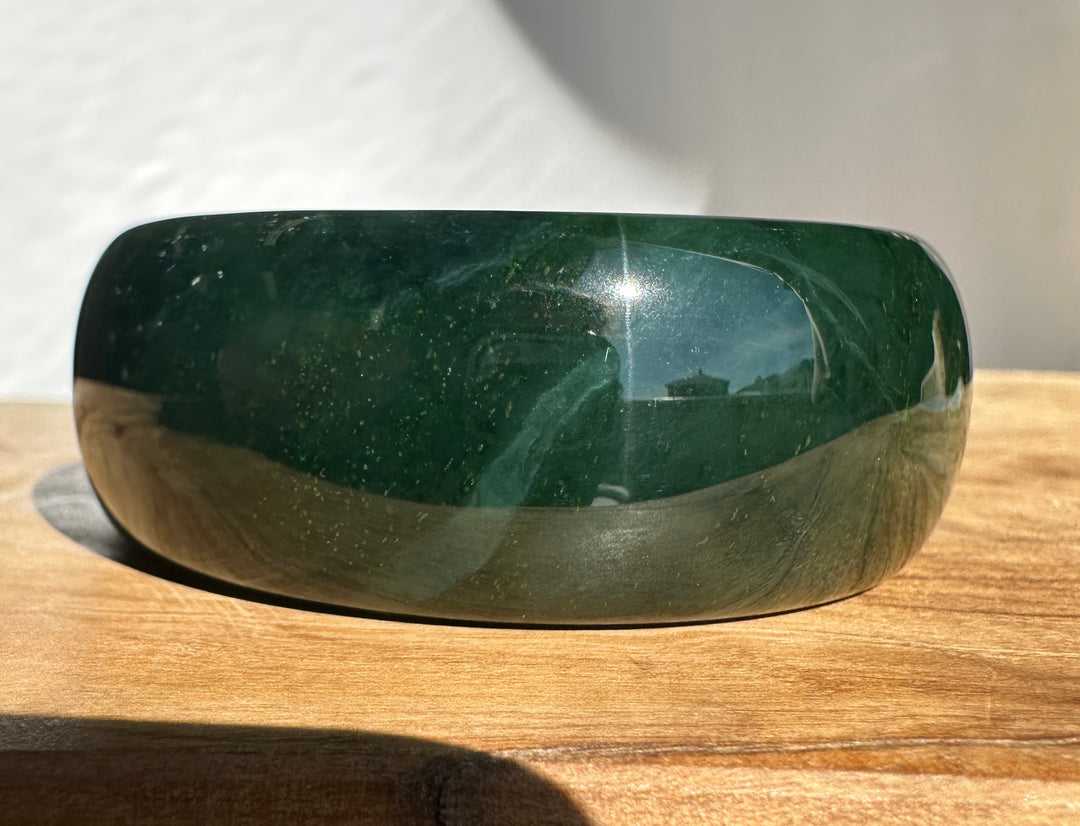 A polished dark green jade bangle with a smooth, rounded shape, resting on a wooden surface under direct sunlight.