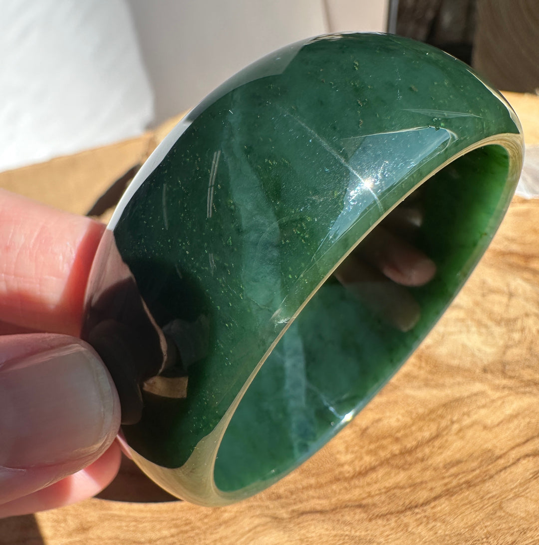 A hand holding a polished dark green jade bangle bracelet against a wooden surface background.