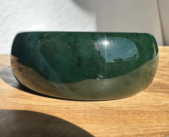 A polished dark green jade bangle with a smooth, rounded shape, resting on a light brown wooden surface under bright natural sunlight.