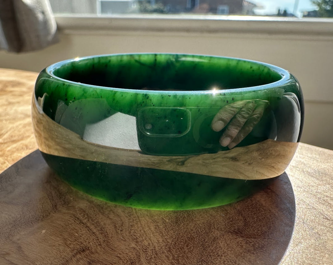 A thick, wide green jade bangle bracelet with a polished finish and a central gold inlay, resting on a wooden table near a window.