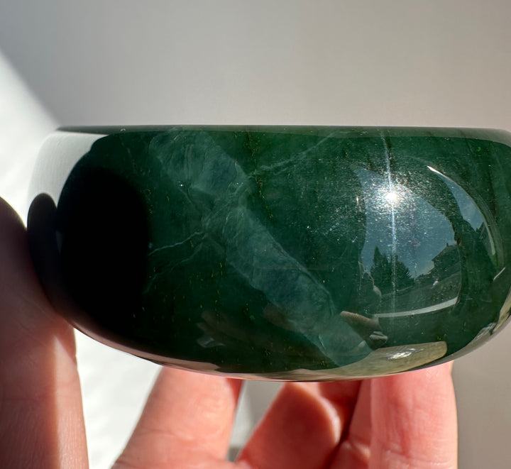 A close-up of a hand holding a polished dark green jade bangle, showing its smooth glossy surface and natural marbled texture.