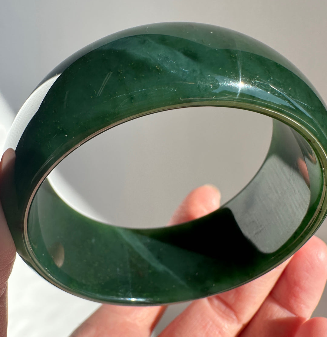 A glossy dark green jade bangle bracelet held in a hand against a neutral light gray background.