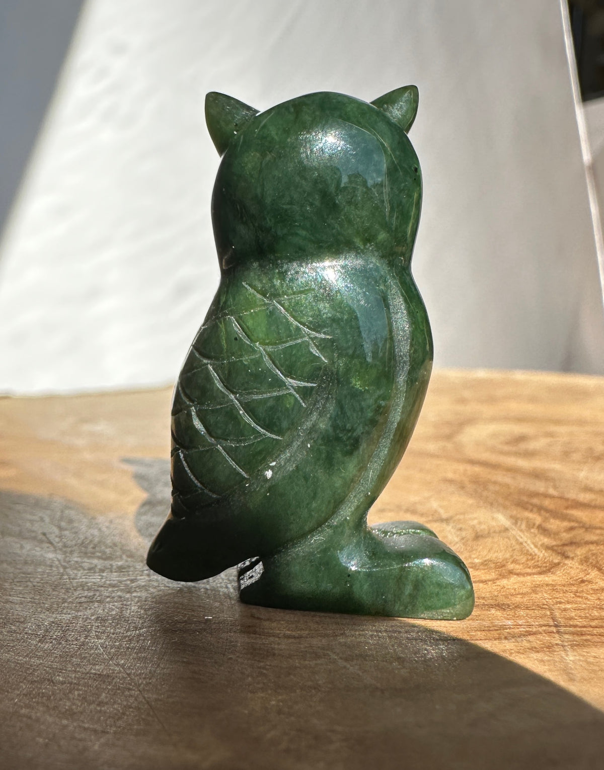 A small carved green stone owl figurine sitting upright on a wooden surface, photographed in natural light against a light neutral background.